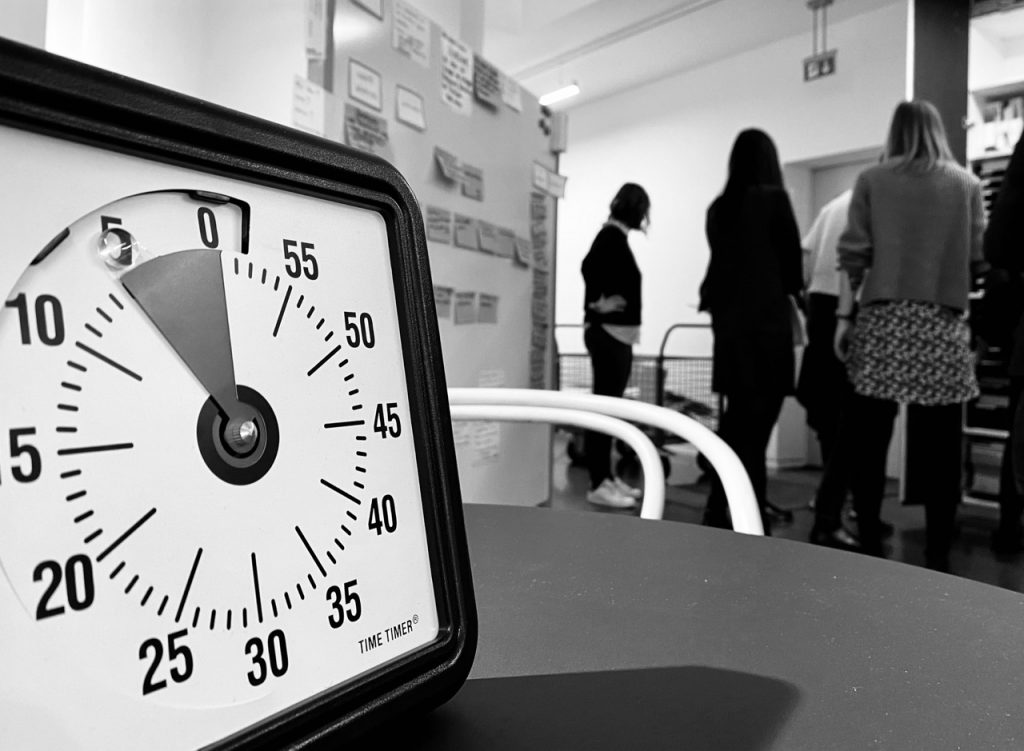 Uhr im Vordergrund mit Workshopteilnehmern im Hintergrund